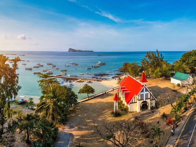 A small church with a red roof on the shore, with boats in the lagoon and an island on the horizon.