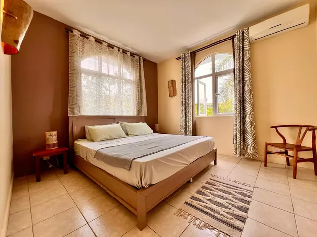 A bedroom with a large bed, a wooden chair and tall curtained windows.