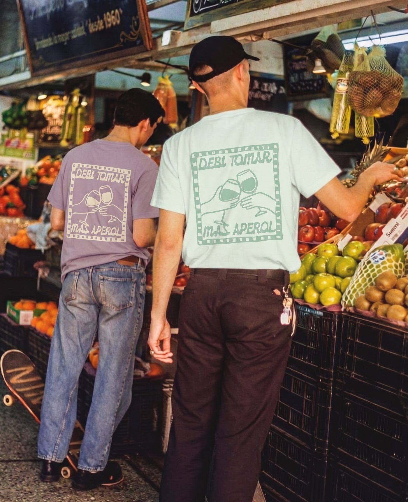 Two friends at a fruit market wearing the lilac and mint Debí tomar más Aperol tees